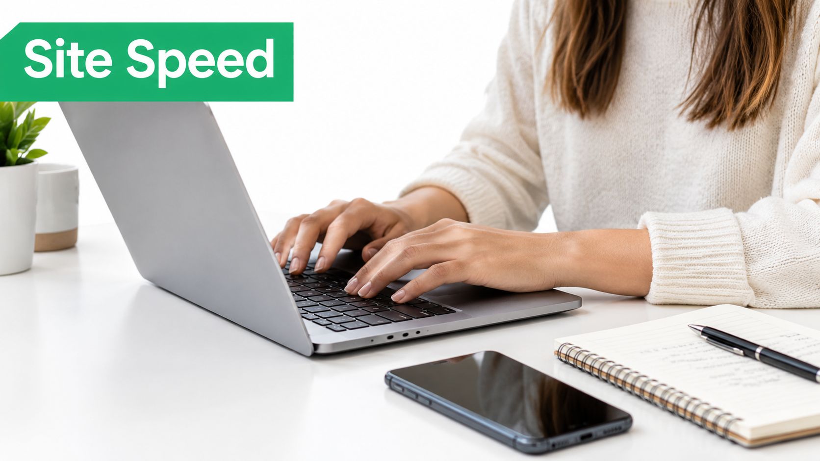 A person wearing a white sweater working on a laptop at a bright desk with a smartphone.